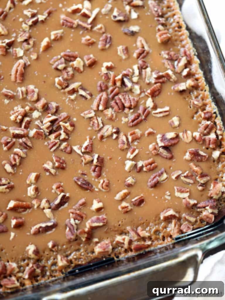 A baking pan filled with freshly baked, uncut caramel pecan brownies, topped generously with golden caramel and chopped pecans. The surface glistens, ready to be sliced into delicious squares.