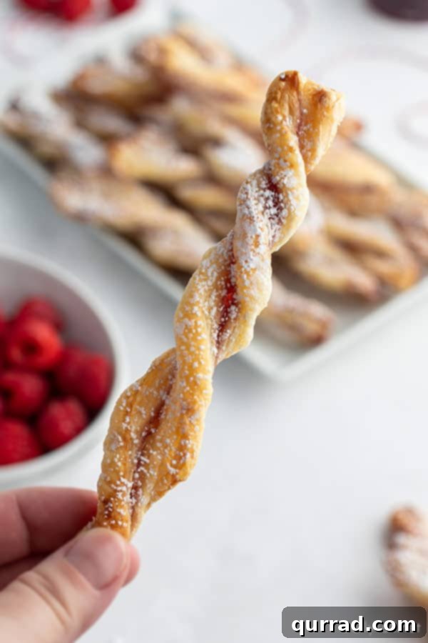Golden Flaky Jam Spirals 6 A close-up shot of a hand delicately holding a single, perfectly baked jam puff pastry twist, showcasing its intricate flaky layers and golden exterior.