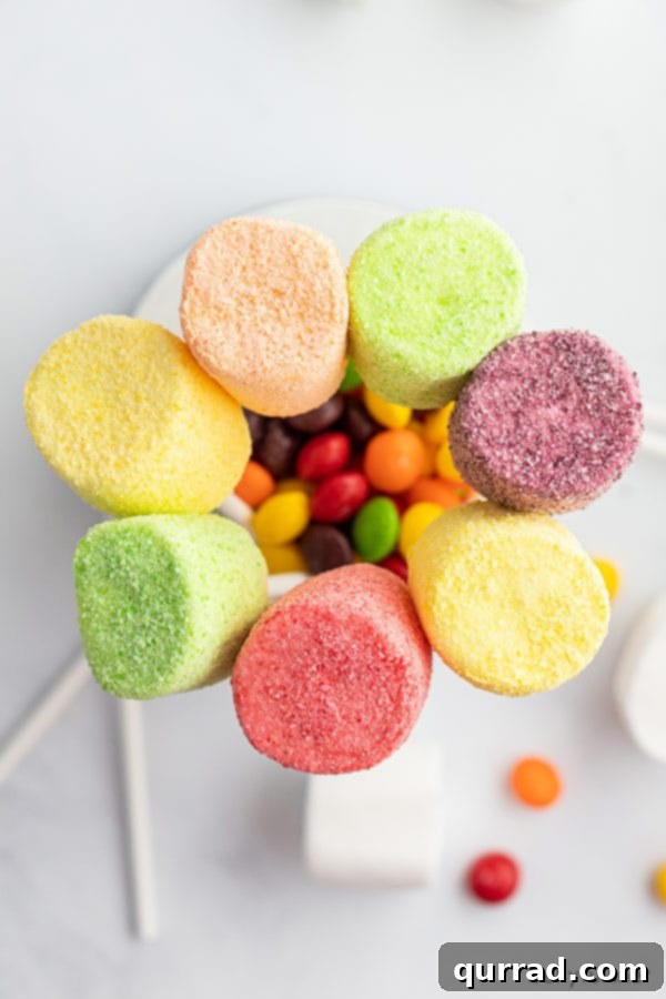 An inviting overhead shot showcasing a variety of rainbow-colored marshmallow pops, each coated in its vibrant Skittles dust, alongside loose Skittles candies.