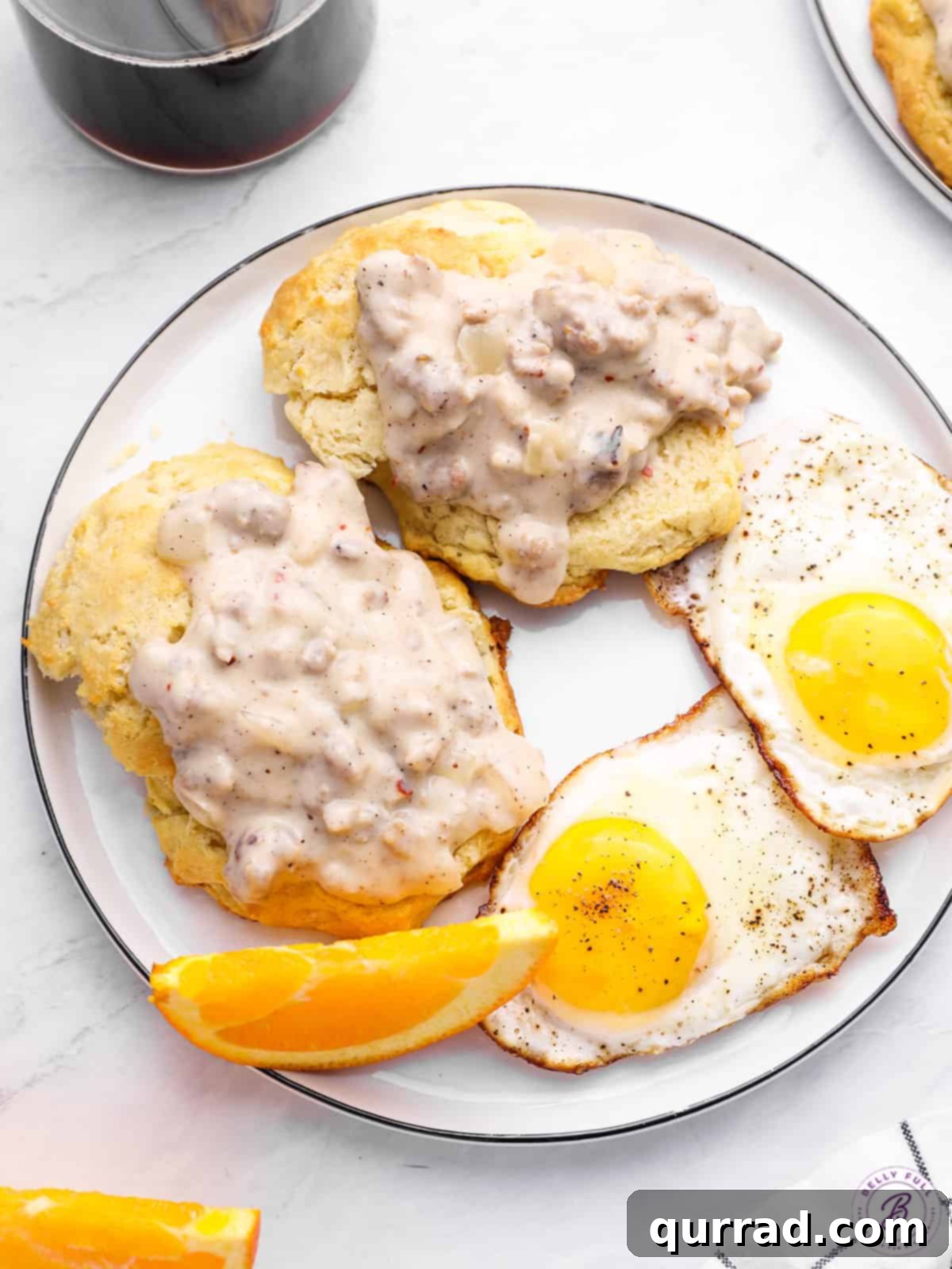 Fluffy Drop Biscuits Smothered in Rich Sausage Gravy 2 Two golden buttermilk biscuits generously covered in creamy sausage gravy, served with two perfectly fried eggs on a white plate, inviting a comforting meal.