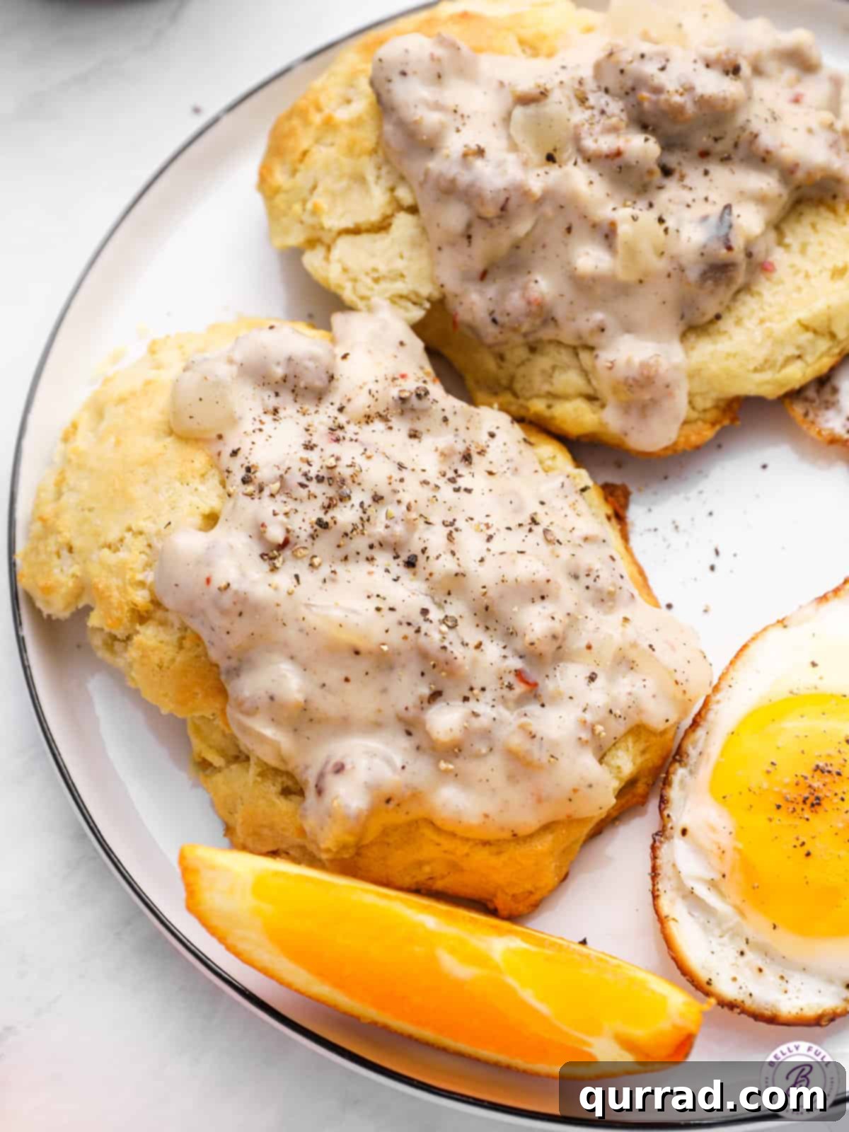Fluffy Drop Biscuits Smothered in Rich Sausage Gravy 8 A close-up shot of golden-brown biscuits generously topped with creamy, spiced sausage gravy, served with a sunny-side-up egg and an orange wedge on a plate.