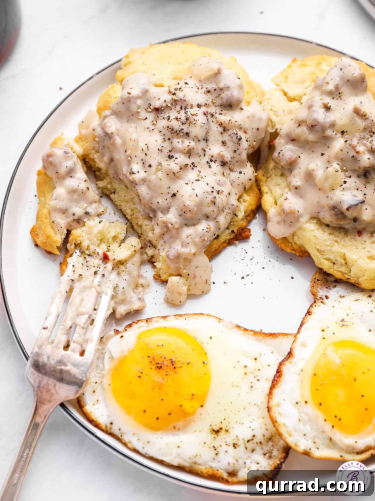 Fluffy Drop Biscuits Smothered in Rich Sausage Gravy 9 A fork lifting a piece of biscuit with gravy and a fried egg from a white plate, highlighting the delicious combination.