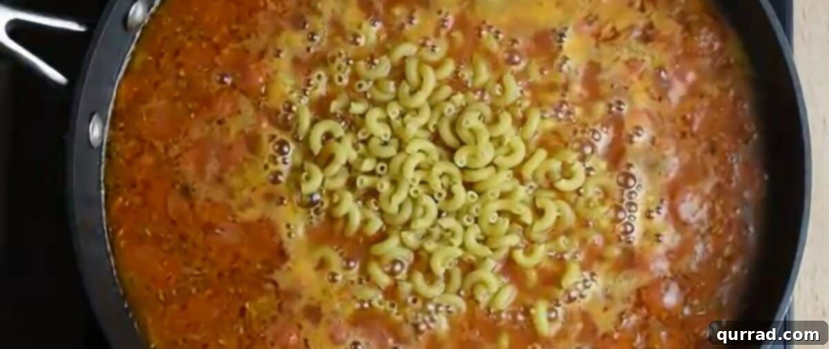 Pizzaroni Mac and Cheese 7 The broth and marinara sauce mixture being poured into a pan with sautéed onions and pepperoni, followed by uncooked macaroni pasta.
