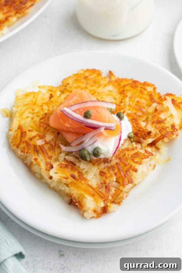 Close-up of a serving of golden hash browns on a white plate, generously topped with slices of smoked salmon, tangy capers, and a swirl of Greek yogurt.