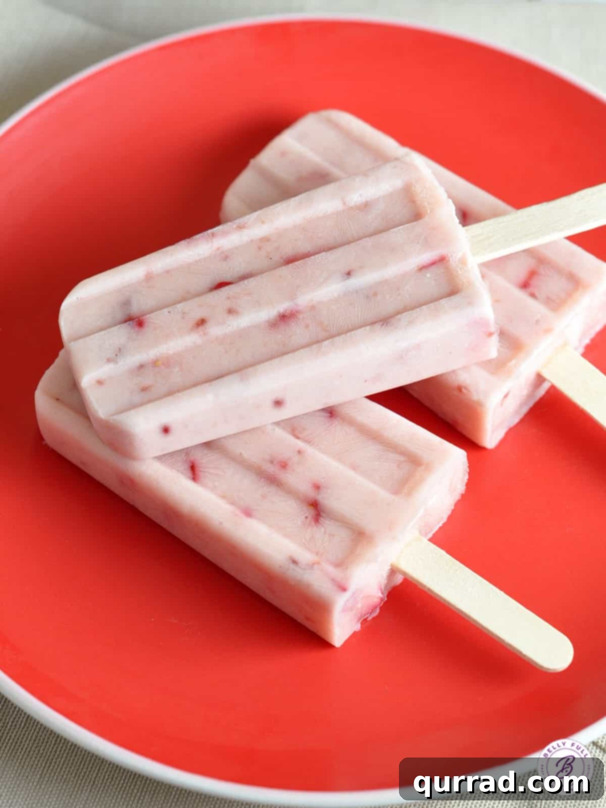 Sweet Strawberry Swirl Yogurt Pops 4 An inviting overhead shot of three creamy strawberry popsicles elegantly arranged on a red plate, highlighting their vibrant color.