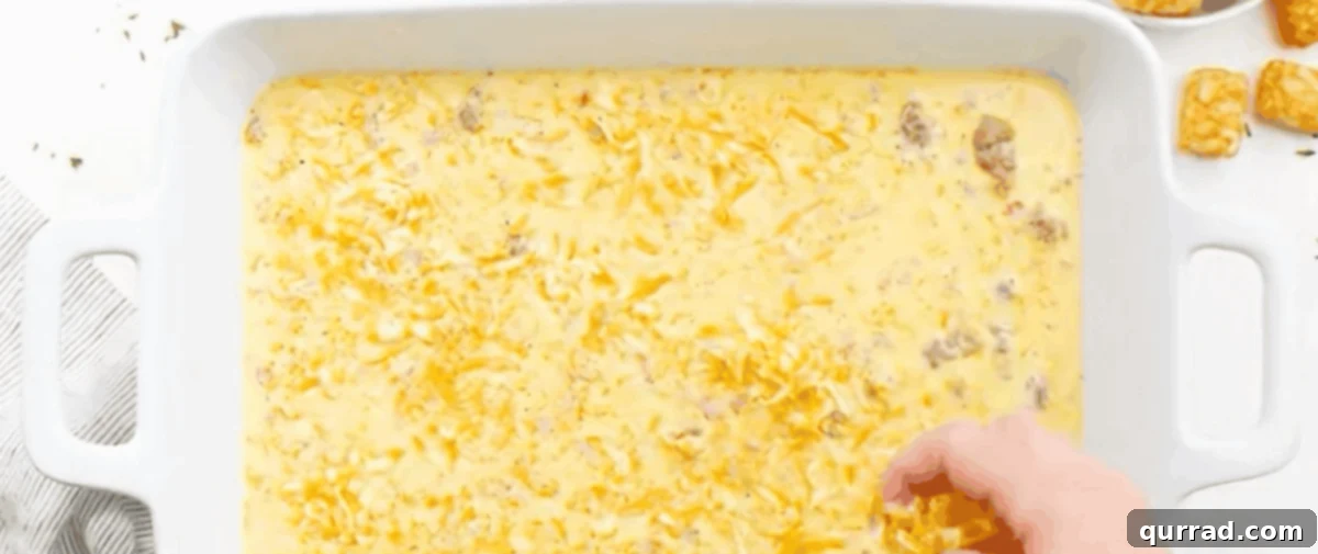 Shredded Colby Jack cheese being sprinkled over the egg and sausage mixture in a baking dish, with tater tots visible.