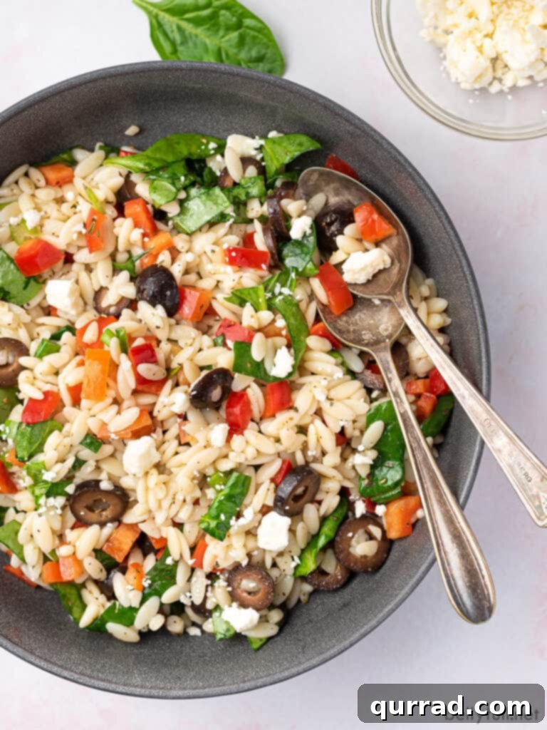 Sun-Kissed Mediterranean Orzo 2 Overhead view of a generous bowl of Mediterranean Orzo Salad, showcasing orzo pasta, olives, red bell pepper, spinach, and crumbled feta cheese, with serving spoons nestled in the bowl.