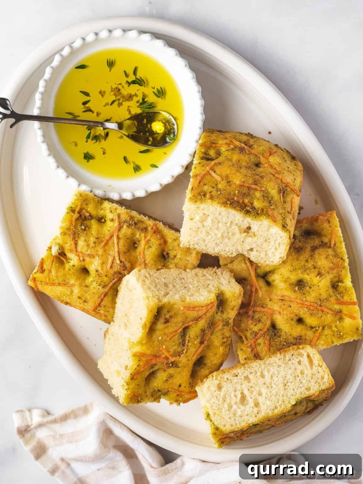 Savory Rosemary Focaccia 2 Golden-brown focaccia bread, generously topped with pesto and Parmesan, cut into squares and arranged on a white serving platter, with a small bowl of olive oil for dipping.
