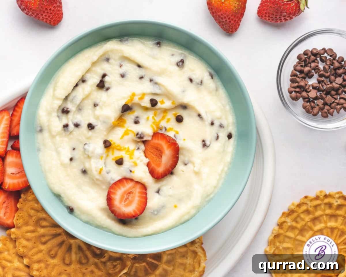 Overhead close-up of a bowl of cannoli dip, perfectly smooth and garnished with mini chocolate chips, surrounded by fresh strawberry halves for dipping.