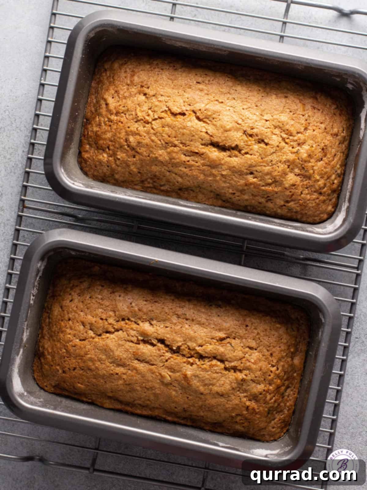 Two golden-brown zucchini bread loaves cooling in their pans on a wire rack after baking.