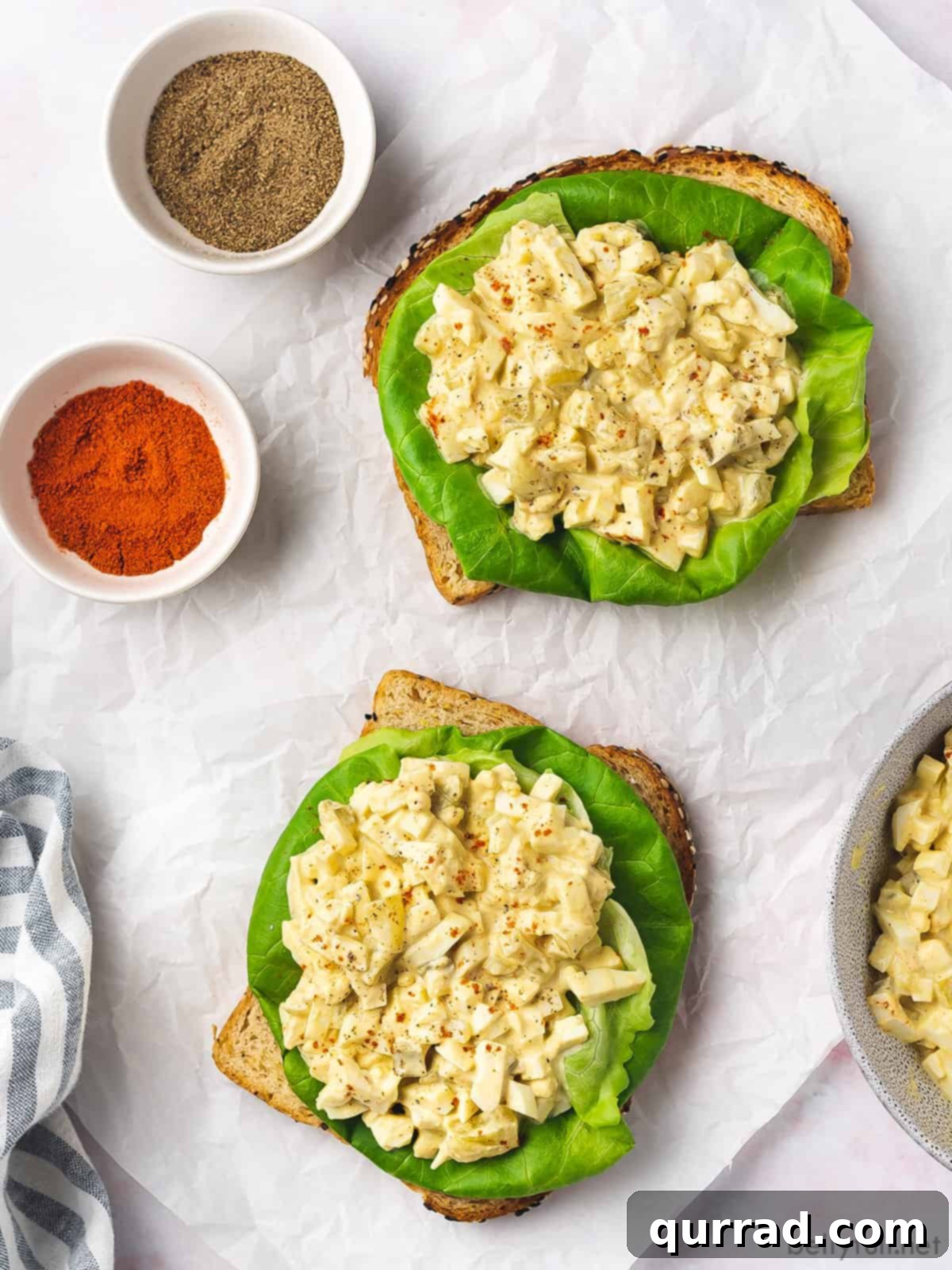 Creamy Egg Salad 8 An appetizing open-face egg salad sandwich garnished with fresh greens and a sprinkle of paprika, served alongside small bowls of black pepper and paprika for seasoning.