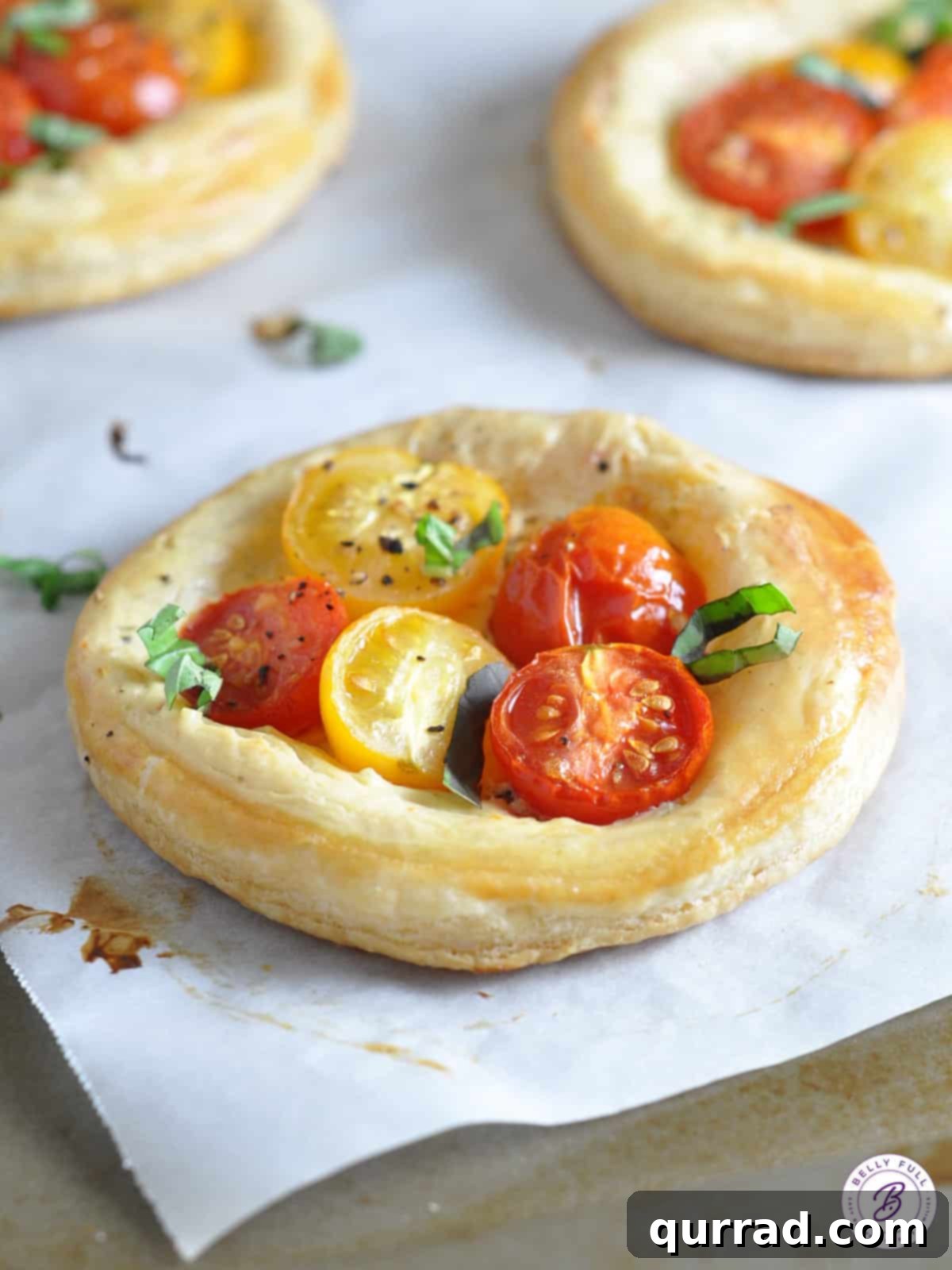 Flaky Tomato Tarts 2 Golden-brown, round individual puff pastry tomato tarts garnished with fresh basil leaves on a white serving plate.