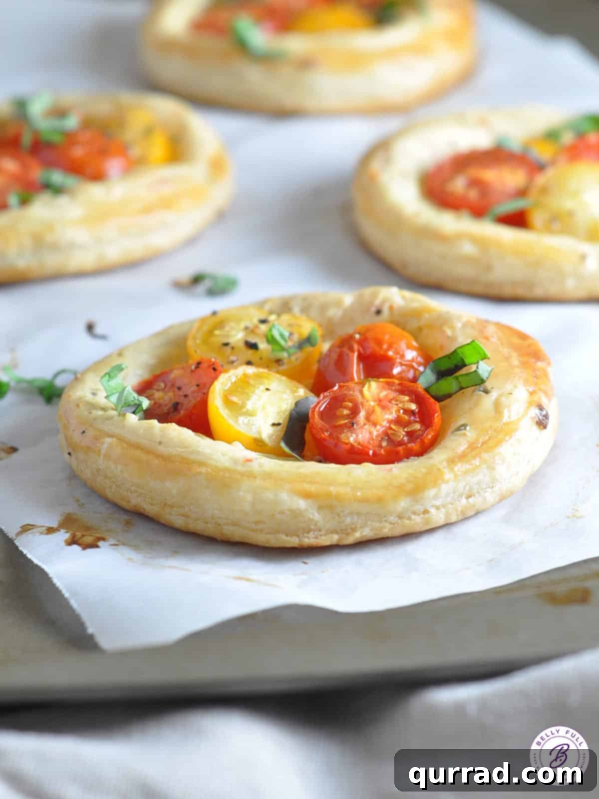 Flaky Tomato Tarts 4 Close-up of a baked individual puff pastry tart with roasted cherry tomatoes and melted cheese, garnished with fresh basil.