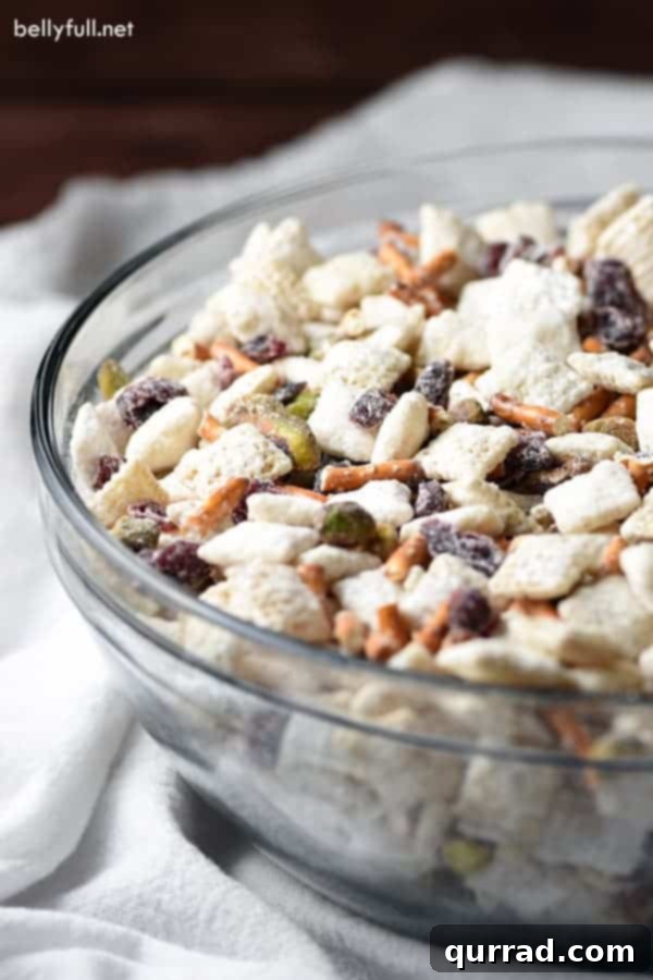 A vibrant glass bowl overflowing with homemade Muddy Buddies, featuring the festive colors of pistachios, red craisins, and crunchy pretzel pieces coated in white chocolate and powdered sugar, ideal for holiday snacking.