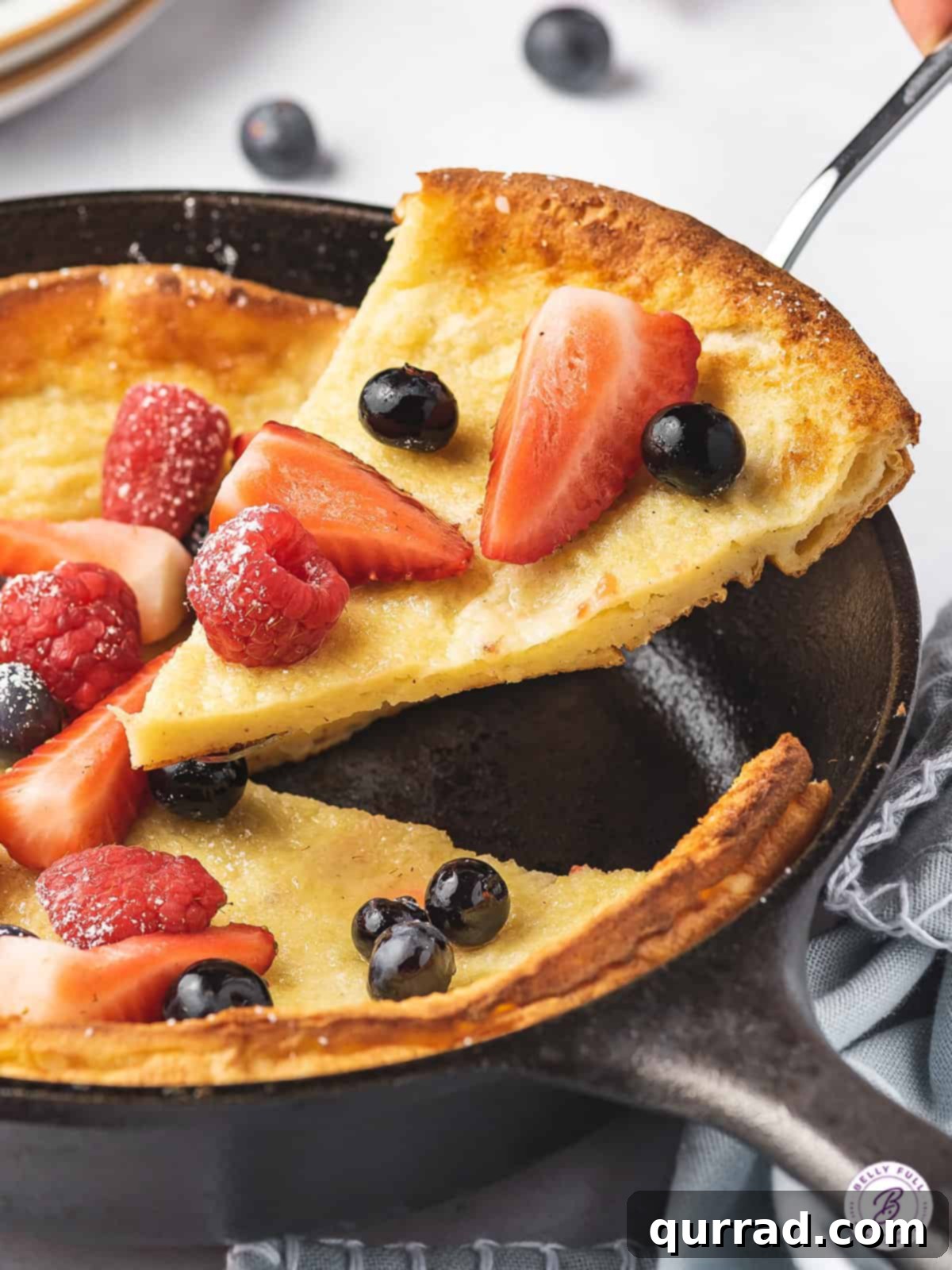 A serving spatula holding a slice of Dutch Baby pancake, garnished with fresh fruit, above the skillet.