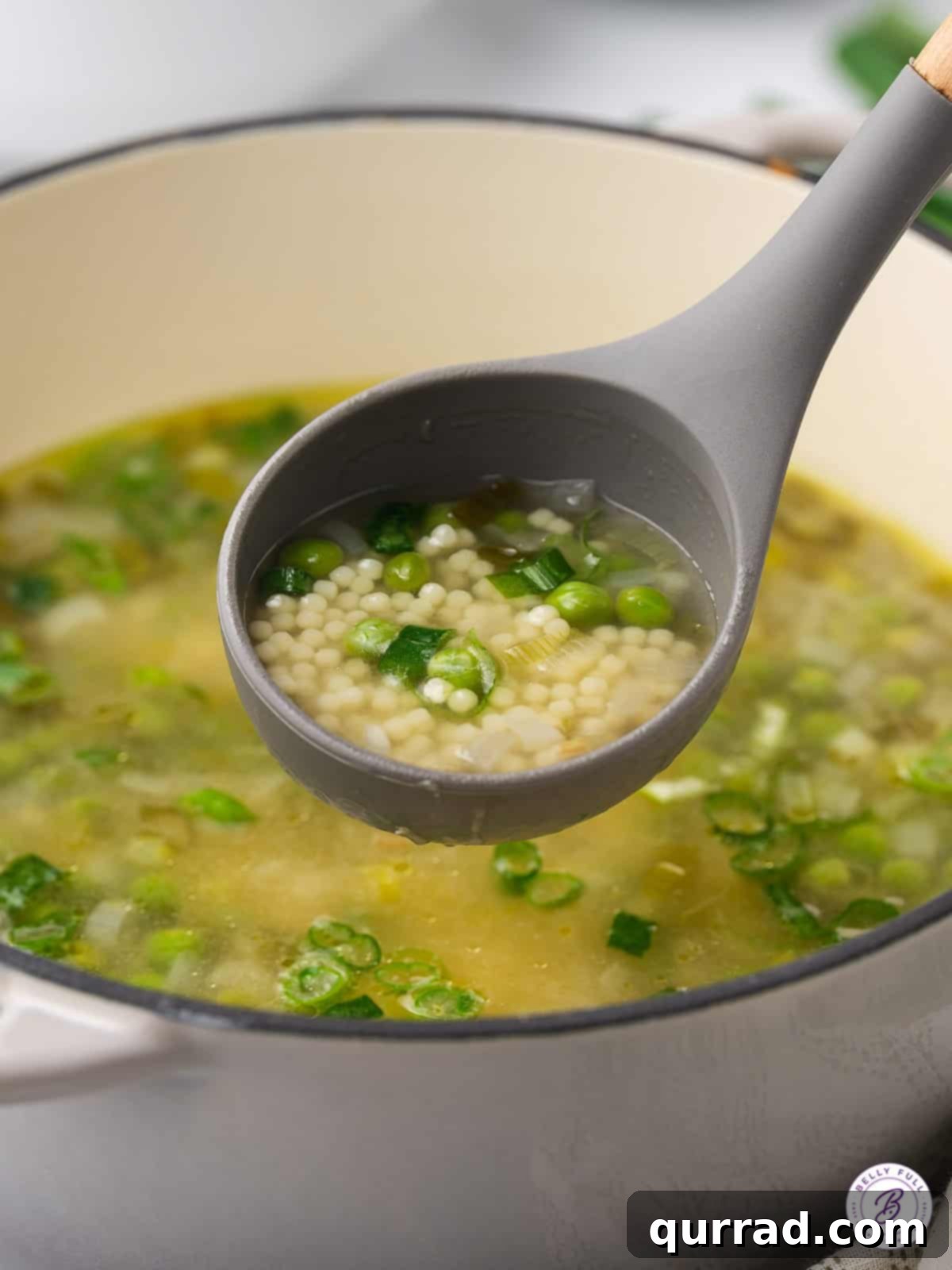 A ladle serving Spring Onion and Pea Soup into a bowl, showcasing the small acini di pepe pasta and vibrant green peas floating in the savory broth.