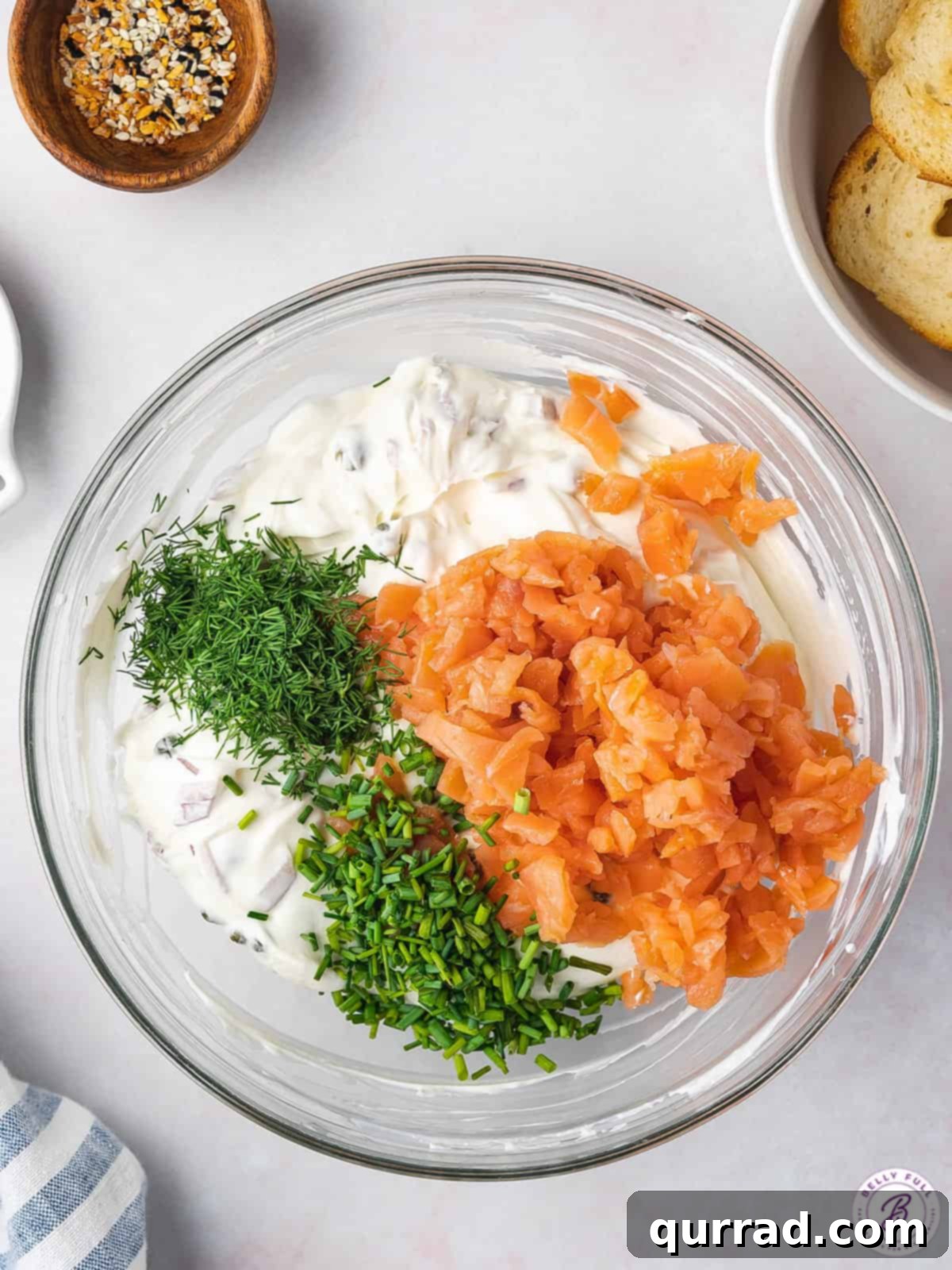 A selection of fresh ingredients laid out in small bowls, including diced red onion, capers, fresh dill, chopped chives, and a block of cream cheese, ready to be combined for the salmon dip.