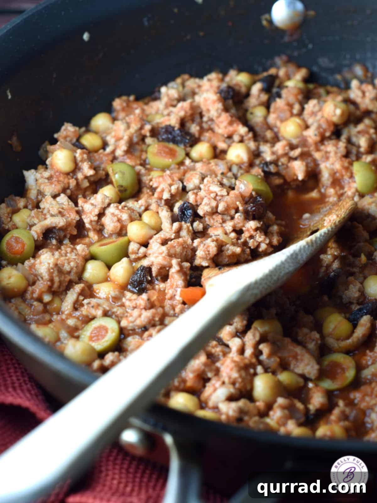 Wooden spoonful of ground turkey picadillo in pan, showcasing the rich sauce and ingredients