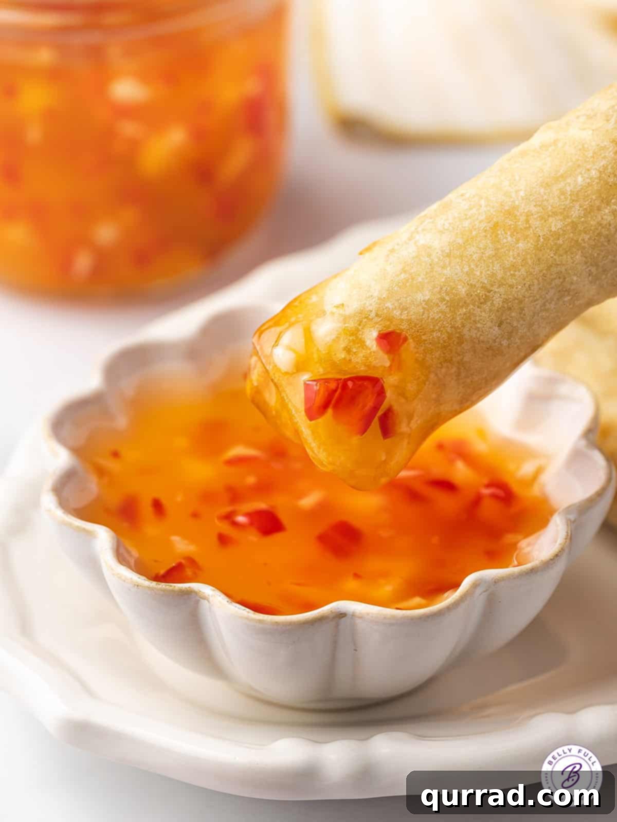 A crispy spring roll being gracefully dunked into a small bowl of homemade Thai sweet chili sauce, highlighting its perfect consistency.