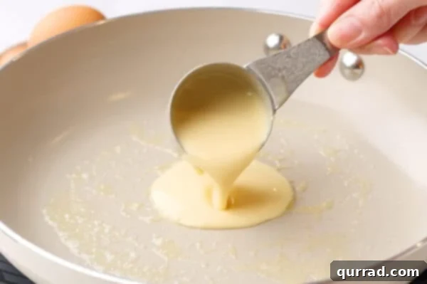Crepe batter being skillfully poured from a ladle into a hot frying pan.
