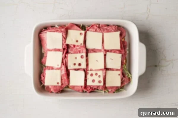 bottom buns in casserole dish with slices of swiss cheese on top of sliced corned beef