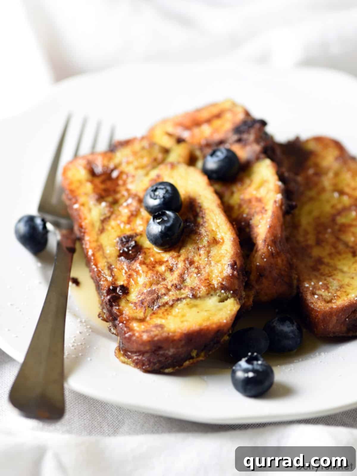 Decadent Chocolate Challah French Toast 3 Slices of golden-brown chocolate challah French toast artfully arranged on a white plate, garnished with vibrant fresh blueberries and a light dusting of powdered sugar.