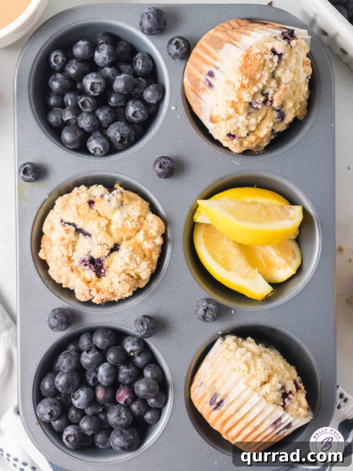 Simple Blueberry Perfection 4 Overhead shot of a 6-slot muffin tin, with three golden-brown blueberry muffins and scattered fresh blueberries.