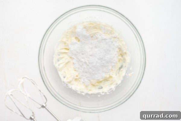 Powdered sugar carefully sifted on top of a whipped butter and cream cheese mixture in a glass bowl, ready for blending.