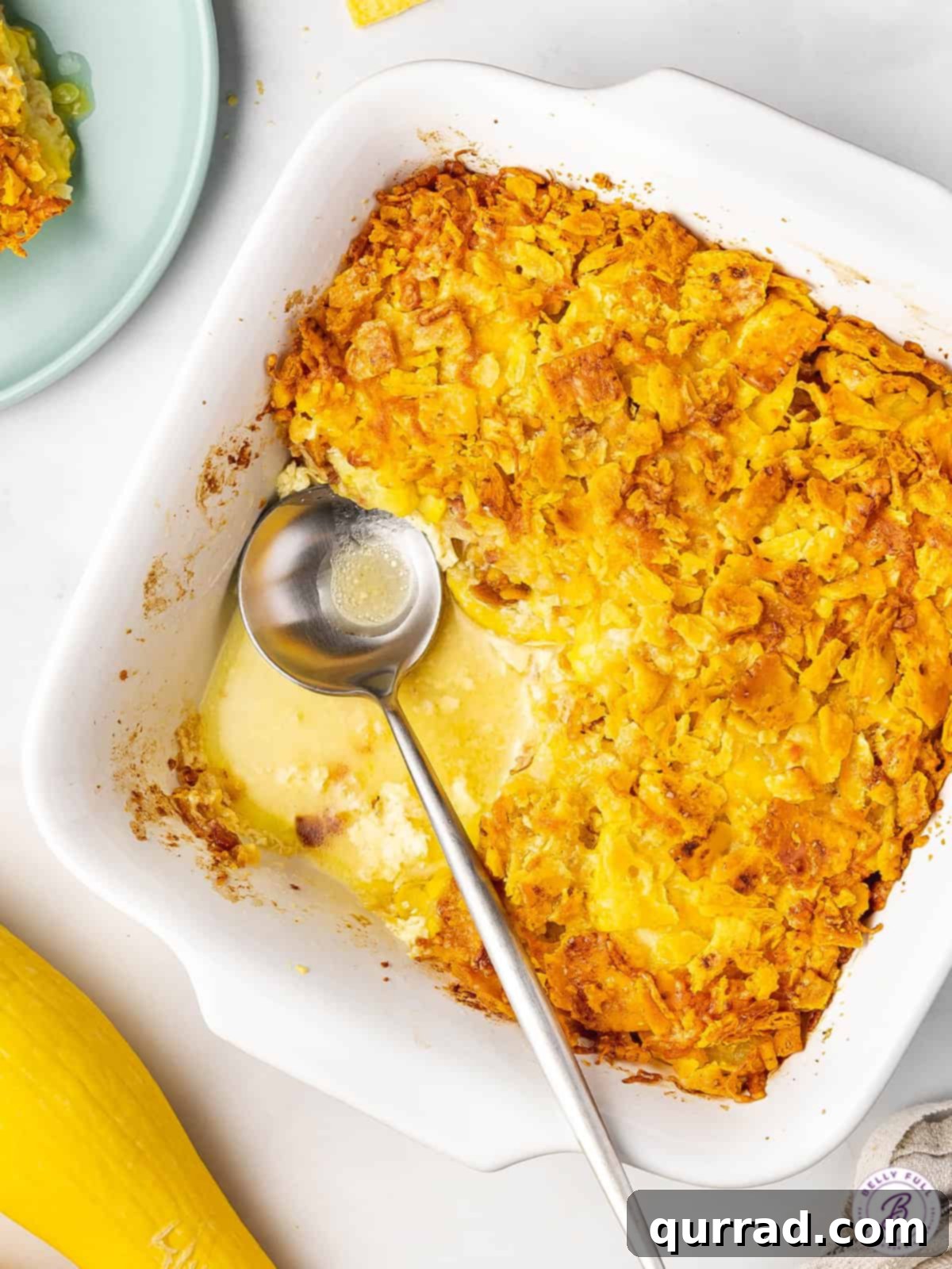 Golden Squash Bake 3 An overhead view of a golden-crusted yellow squash casserole in a white baking dish, with a serving spoon tucked into the side.