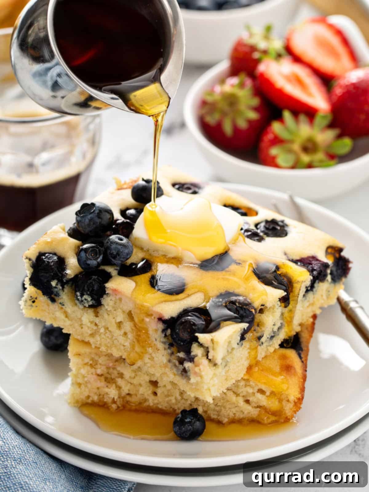 Two square blueberry pancakes stacked with maple syrup being poured on top, ready to be enjoyed.