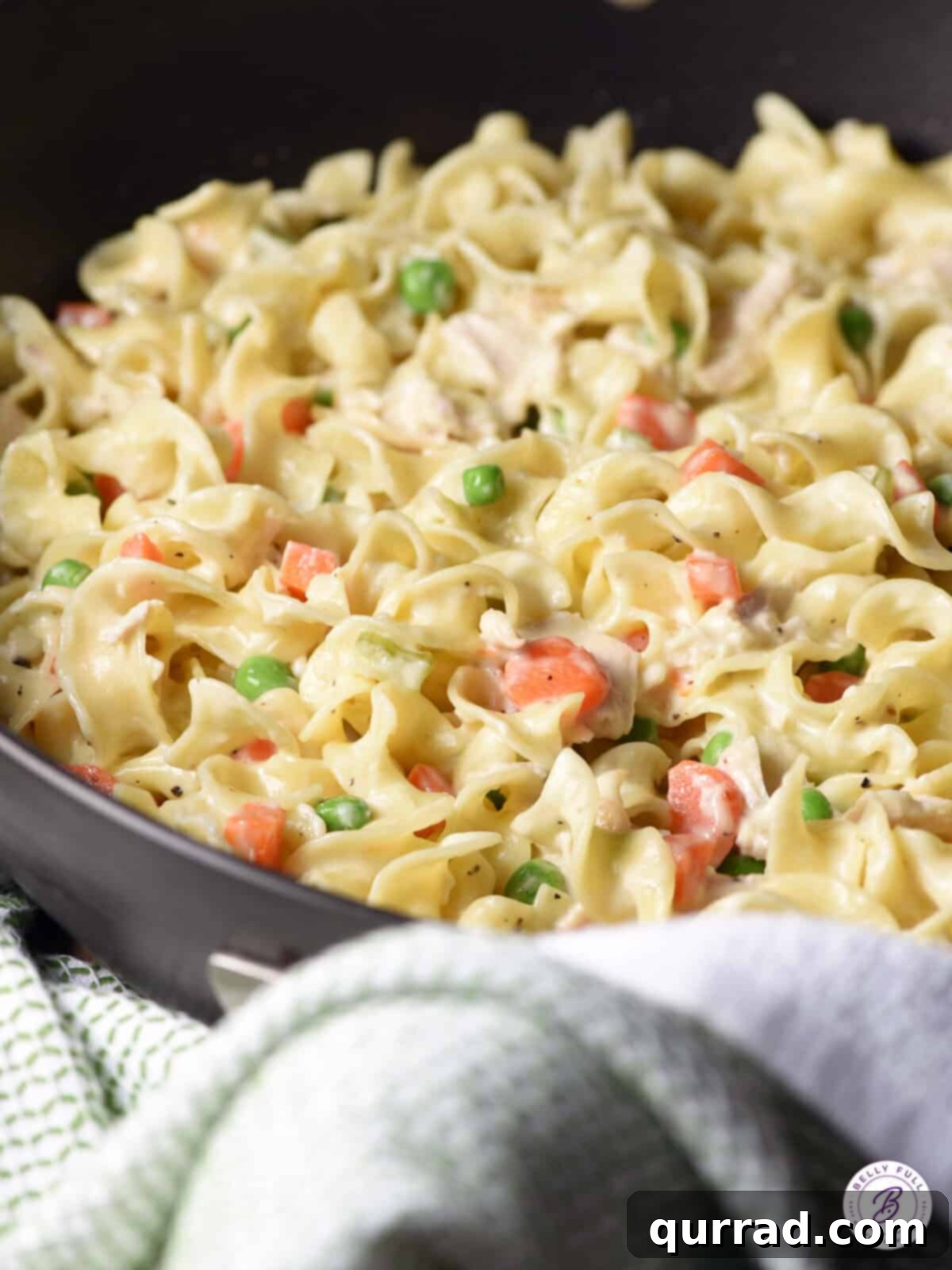 Close-up of a skillet filled with the finished chicken pot pie noodles, showing the rich, creamy sauce coating every strand of egg noodle, along with generous pieces of chicken, peas, and carrots. A true comfort food delight.