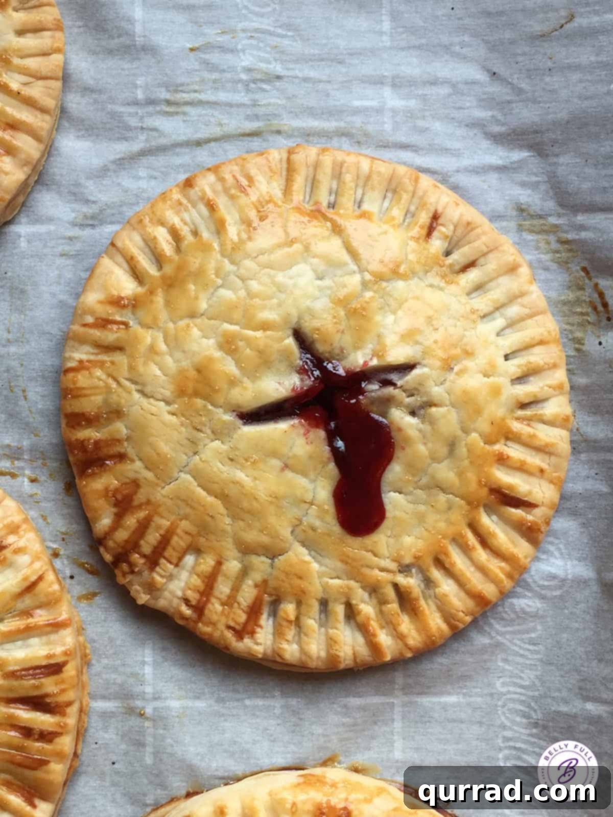 An overhead shot of a freshly baked cranberry hand pie, beautifully glazed, resting on parchment paper. The pie exhibits a golden, flaky crust and a hint of the crimson filling peeking through.