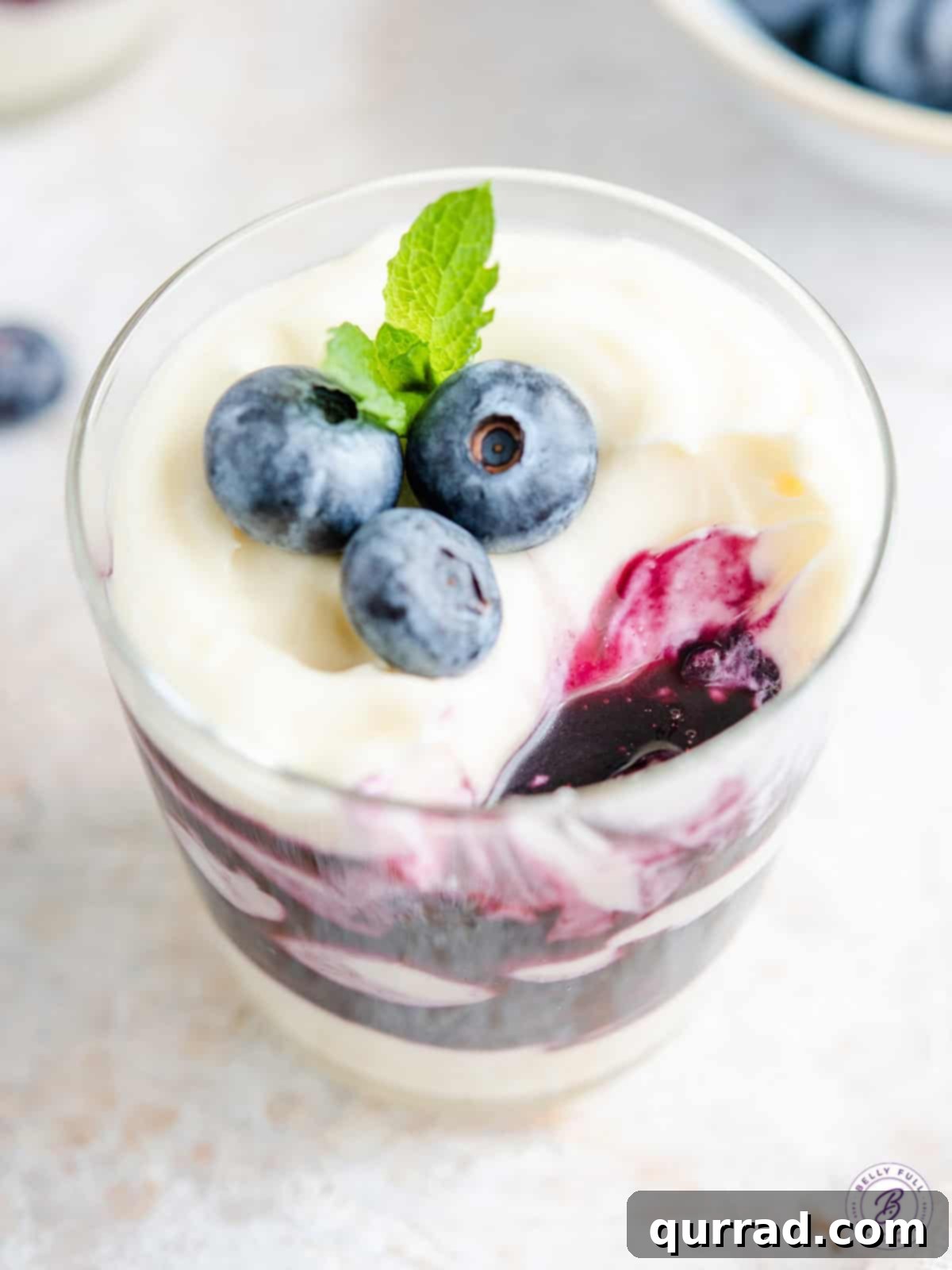 Two glass jars filled with layered blueberry cheesecake parfaits, topped with fresh whole blueberries and a mint sprig on a rustic wooden surface.