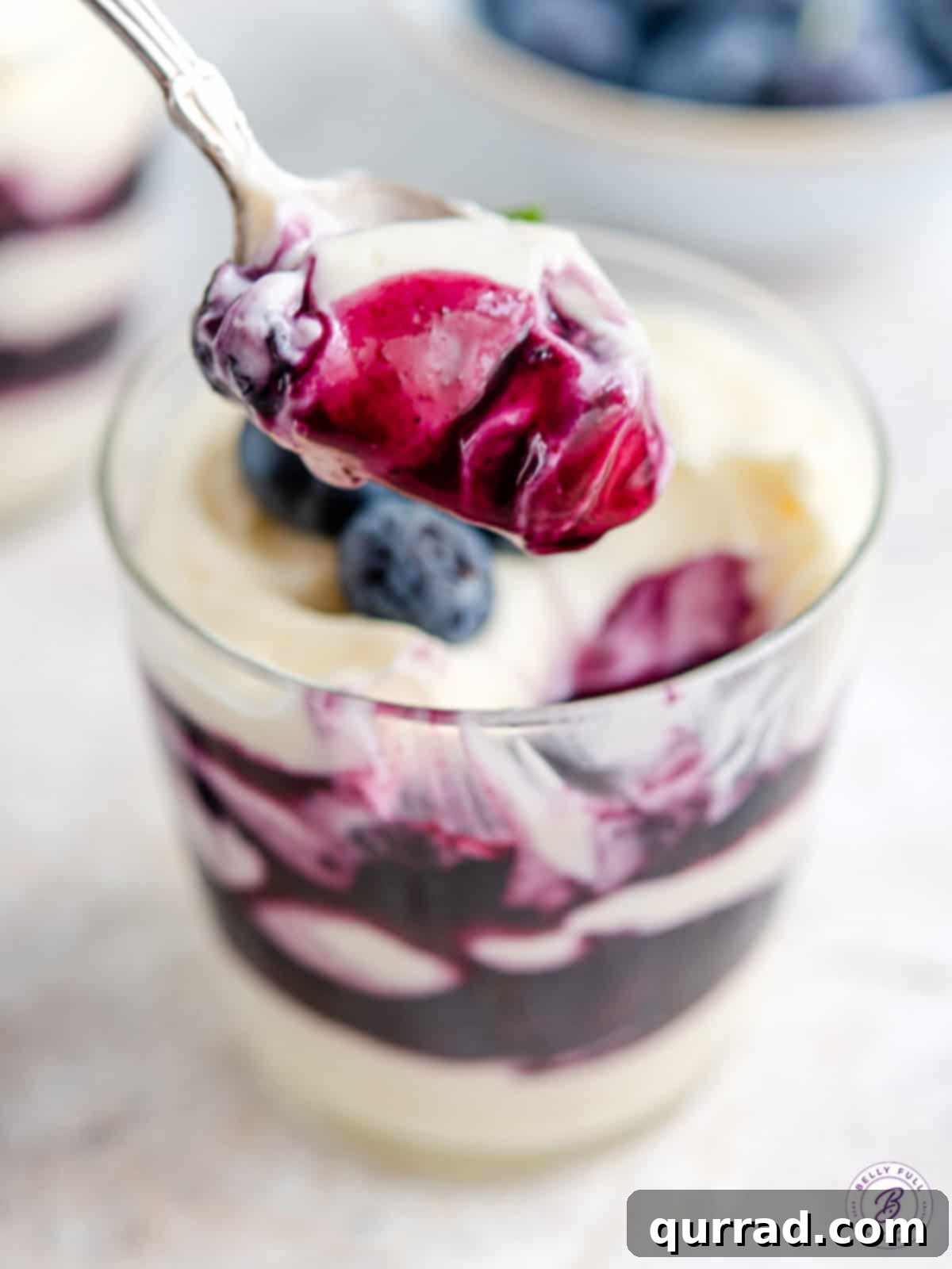 A spoon scooping a portion of blueberry cheesecake parfait, showing the creamy cheesecake and chunky blueberry sauce textures.