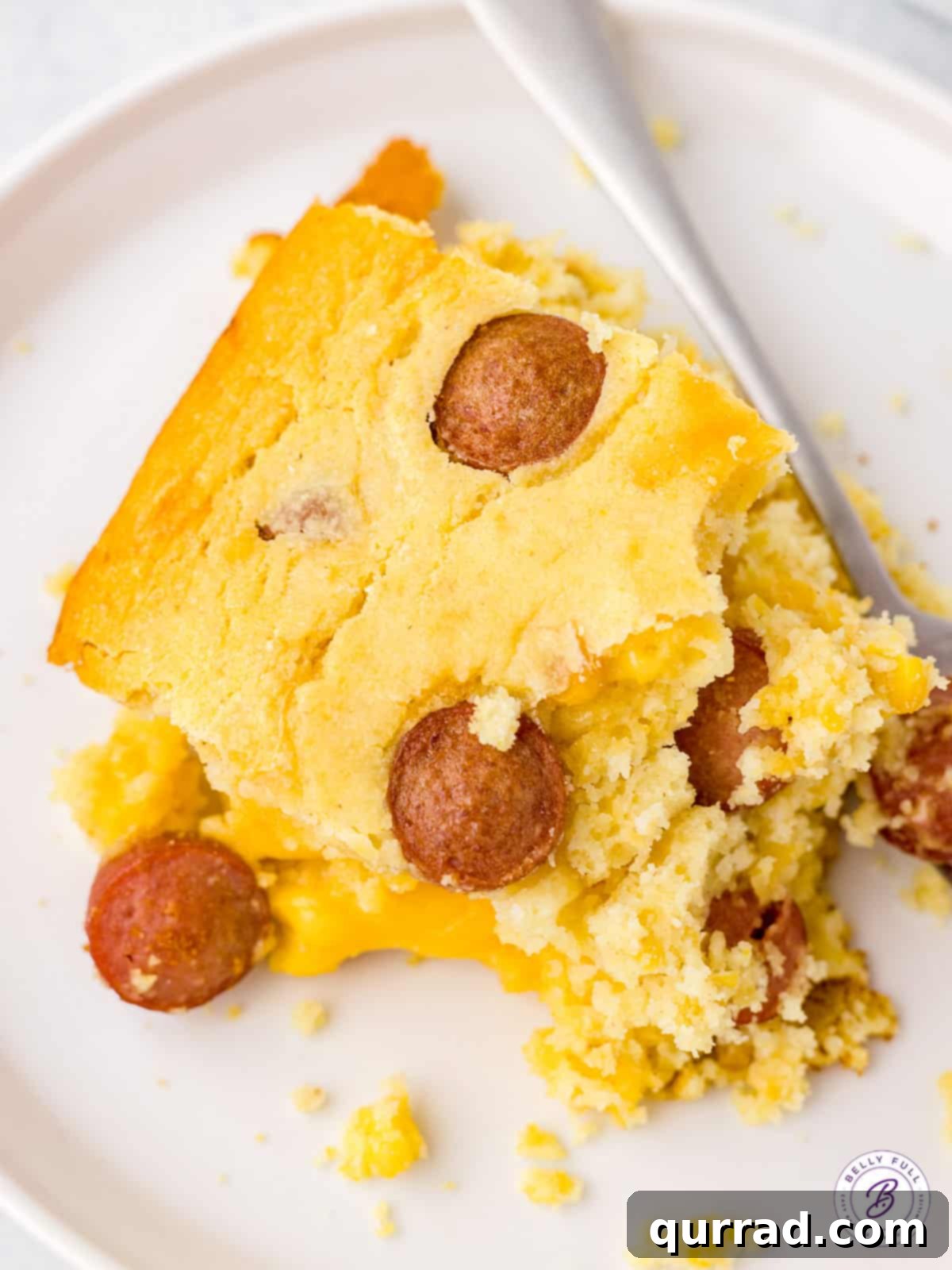 A square piece of golden brown Corn Dog Casserole served on a white plate, showcasing the hot dog slices and cheesy cornbread.