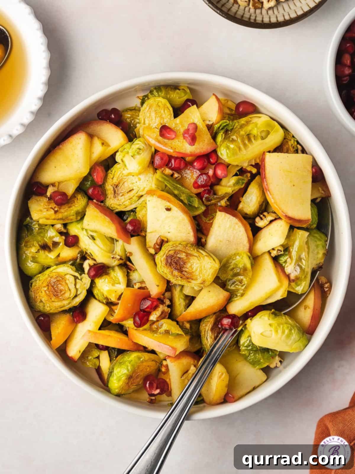 Sweet & Savory Roasted Brussels with Apples 4 Close-up of cooked brussels sprouts with apples and pomegranate seeds in a bowl with a serving spoon.