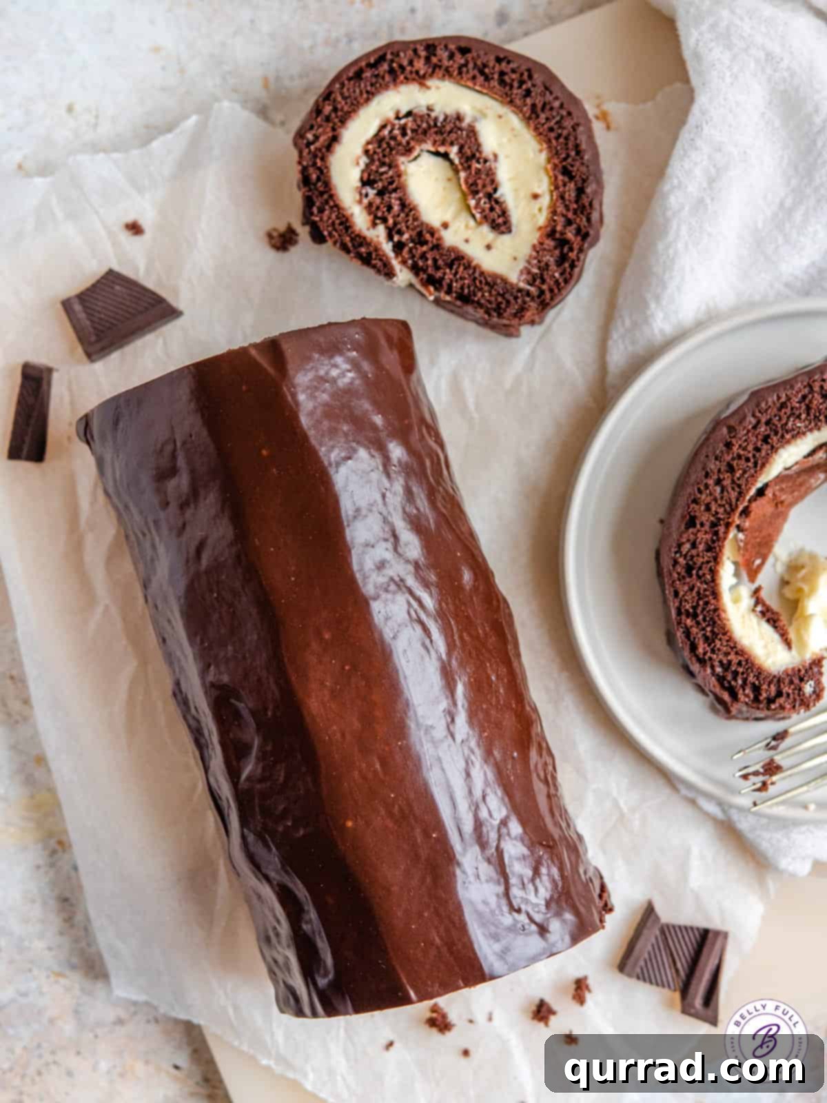 This easy Chocolate Swiss Roll Cake has a light sponge, creamy filling, and coated with a silky ganache. Whole chocolate Swiss roll with a slice on a plate and a slice on parchment paper.