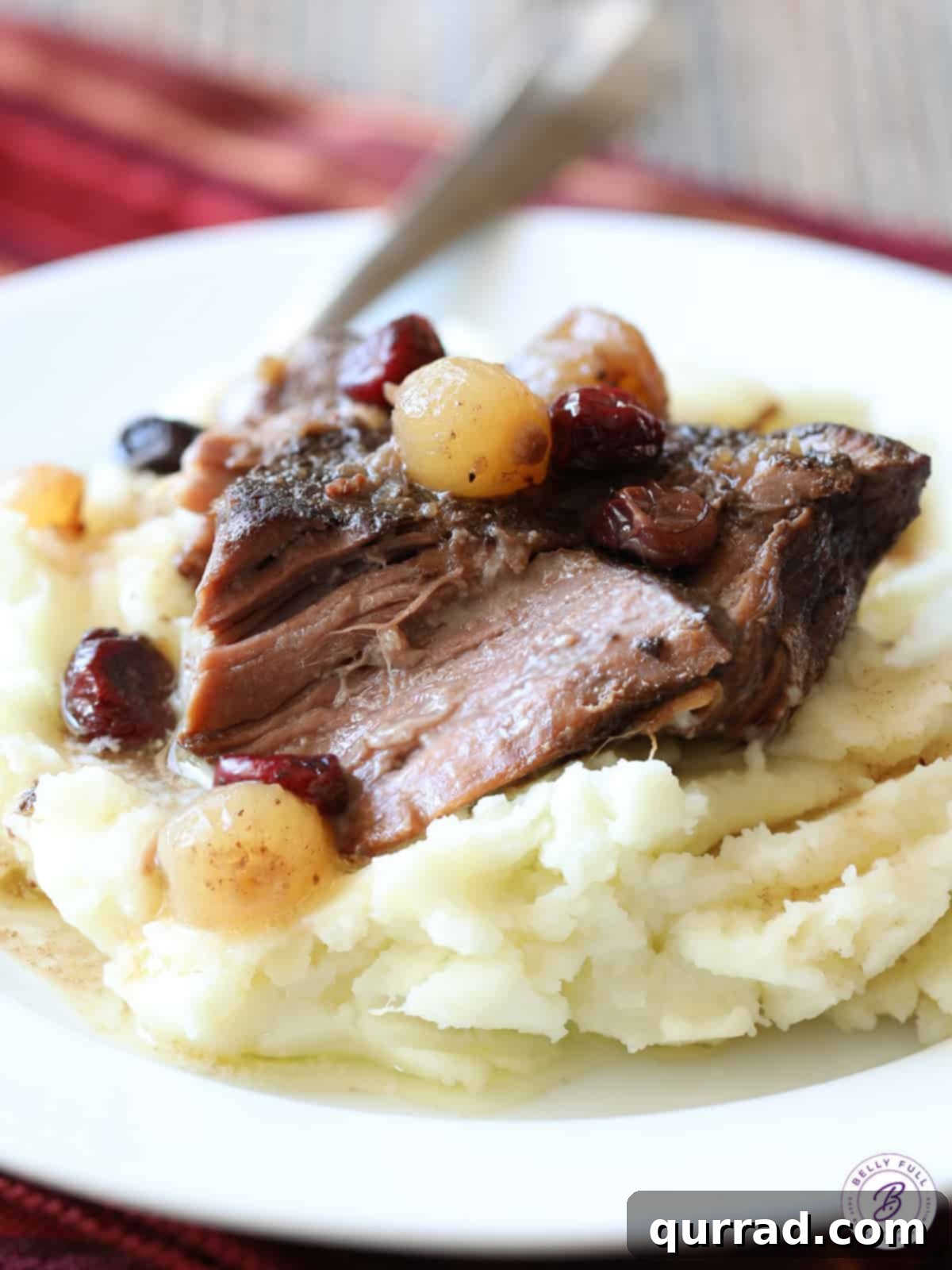 Slab of perfectly cooked pot roast with cranberries and pearl onions served over creamy mashed potatoes, garnished with fresh herbs