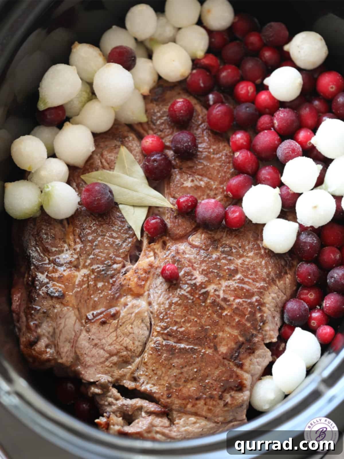 Chuck roast, frozen cranberries, and pearl onions inside a slow cooker before cooking, arranged for maximum flavor infusion
