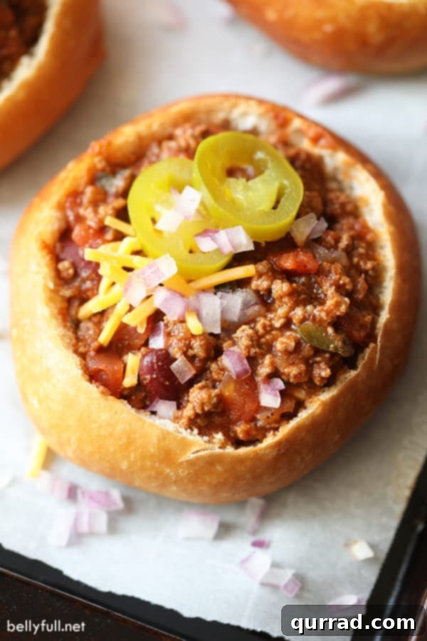 Mini bread bowls filled with chili, topped with sliced pickled jalapenos, diced red onion, and shredded cheese, ready to be served on a rustic wooden board.