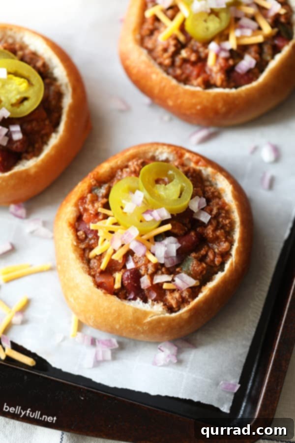 A close-up, top-down view of a mini bread bowl filled with rich, red chili, adorned with vibrant green pickled jalapeno slices, highlighting its appealing presentation.