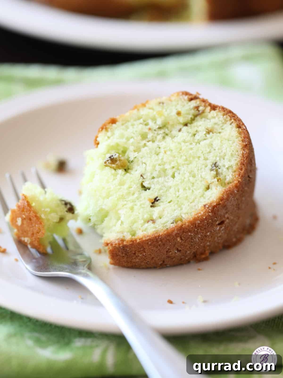 A close-up of a single slice of Pistachio Cake served on a white plate with a fork, emphasizing its tender, moist crumb and vibrant green color.