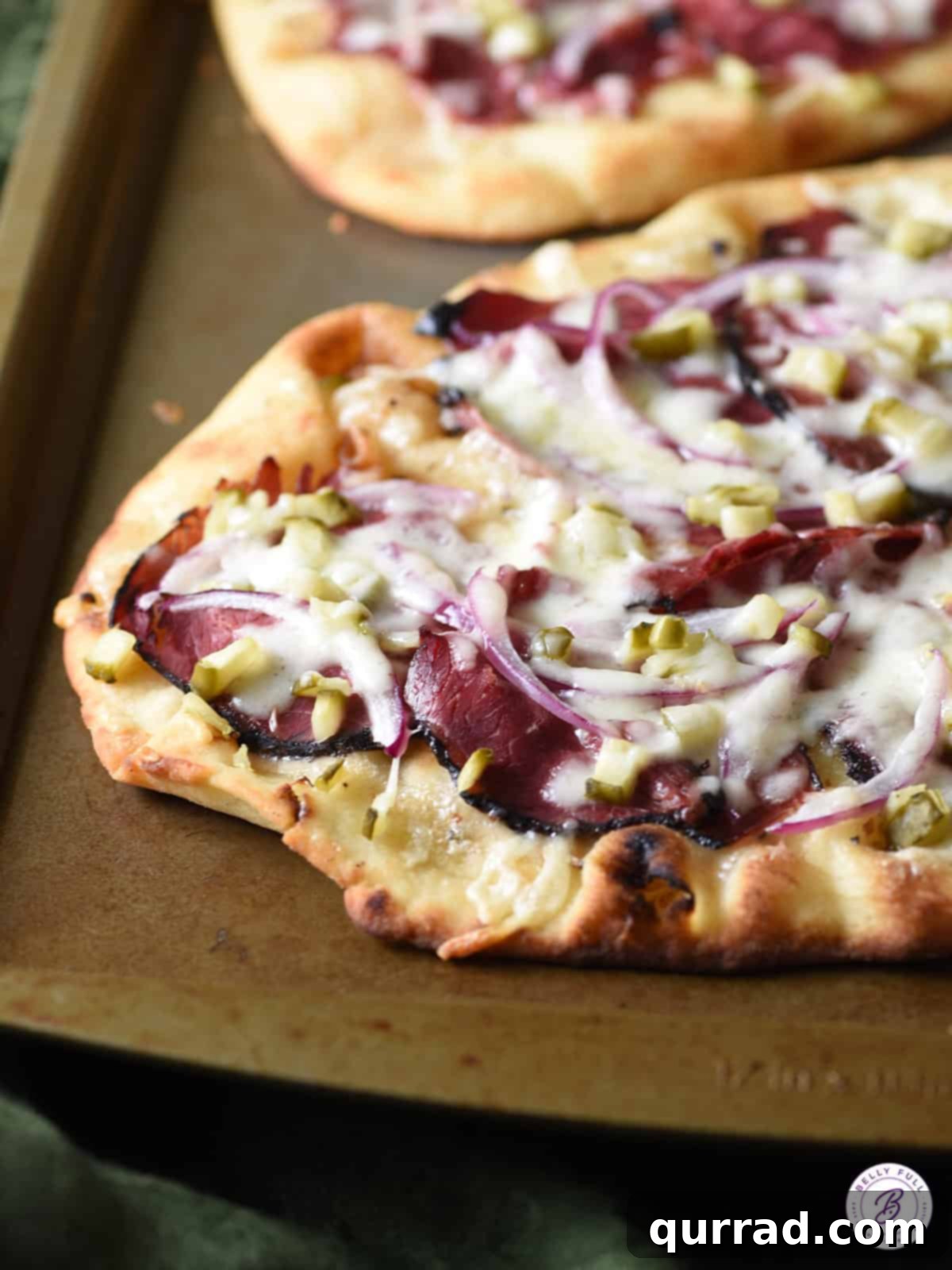 Close-up shot of a freshly baked flatbread with bubbly melted Swiss cheese over pastrami, red onions, and dill pickles, glistening with Caesar dressing.