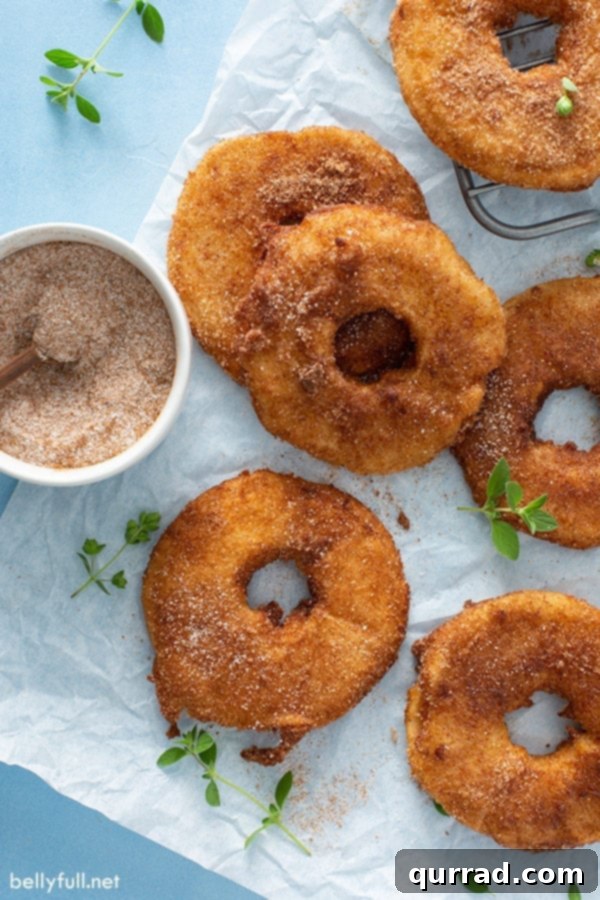 Tropical Golden Fritter Rings 4 Overhead view of freshly fried pineapple fritter rings cooling on parchment paper, next to a bowl of cinnamon-sugar for coating.