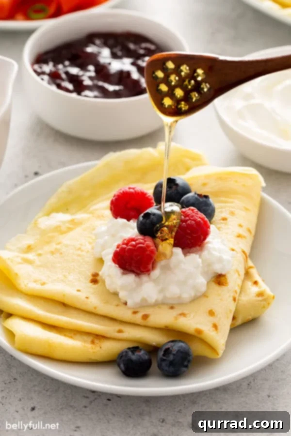 Authentic Russian Blini Recipe 4 2 folded crepes on white plate with cottage cheese, fresh berries, and a drizzle of honey