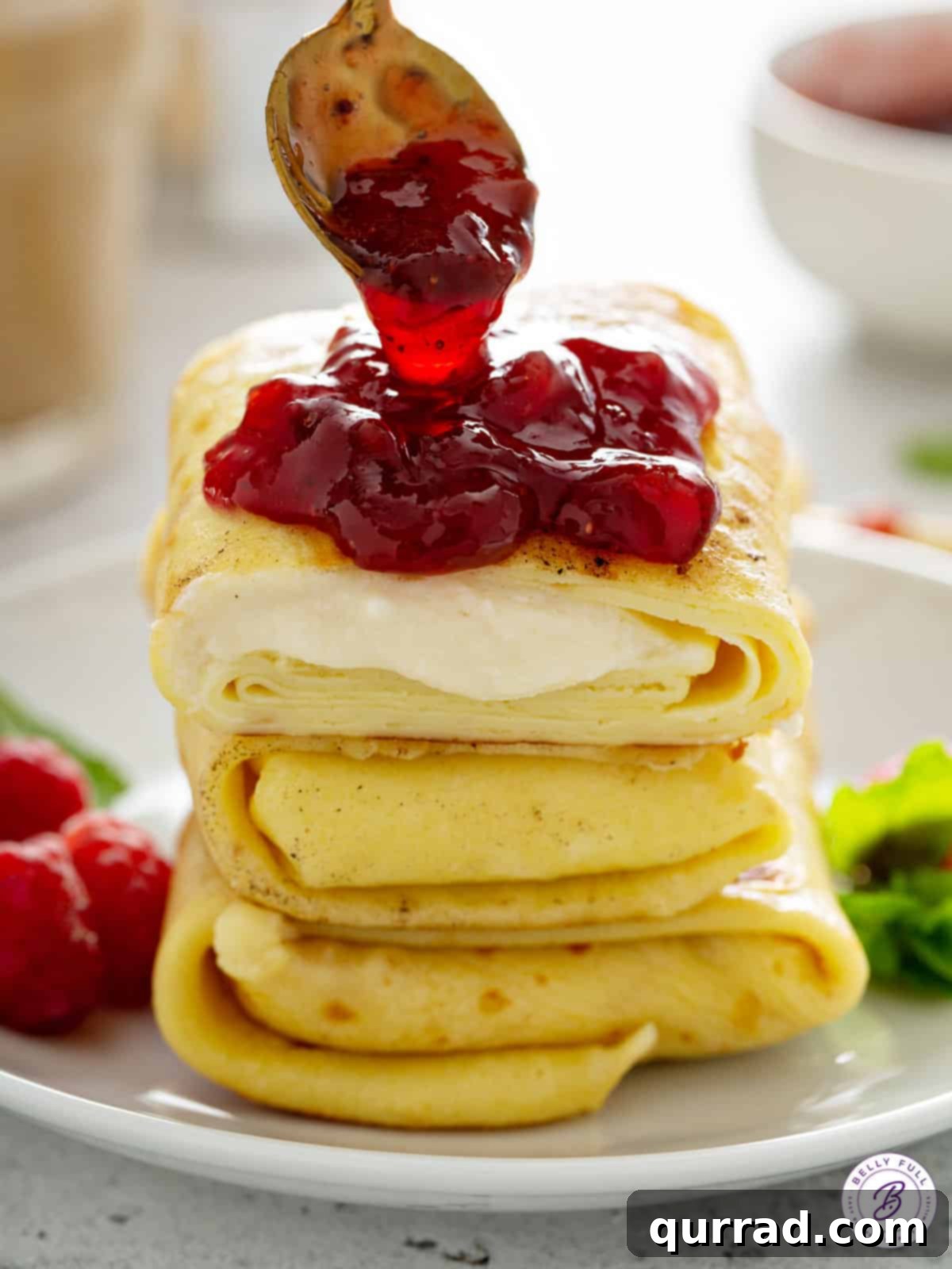 Blintz Bliss 14 A hand spooning bright red jam onto a stack of three freshly prepared, golden-brown blintzes.