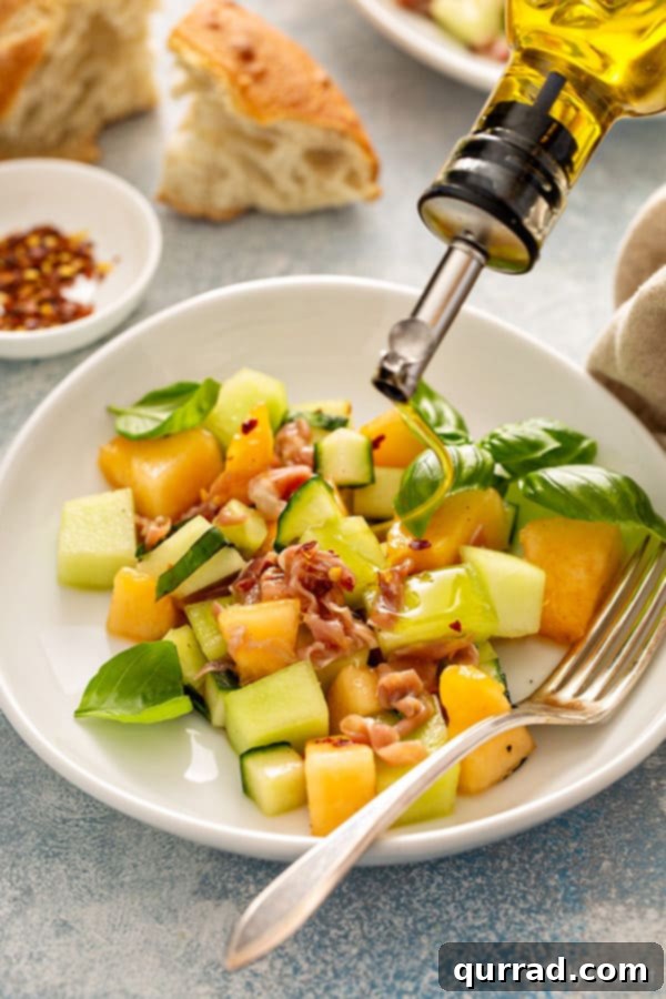 Close-up of Melon Cucumber Salad on a white plate with a fork, and an olive oil bottle drizzling oil on top.