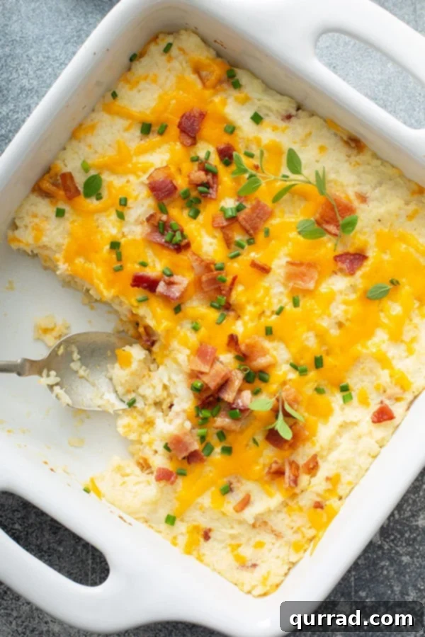Ultimate Loaded Cauliflower Bake 2 Overhead view of a creamy mashed cauliflower bake in a white baking dish with a serving spoon, garnished with crispy bacon and fresh chives.