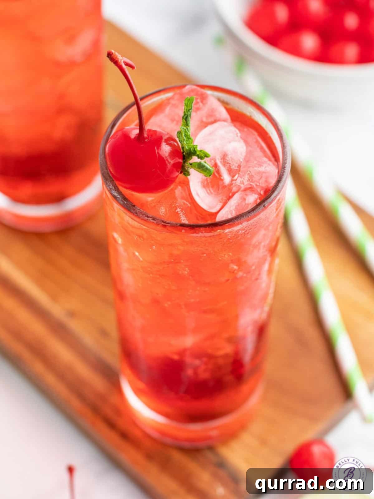 Close-up of a sparkling pink Shirley Temple drink, featuring glistening ice cubes, a bright red maraschino cherry, and a hint of fresh mint on a vibrant background, highlighting its refreshing appeal.