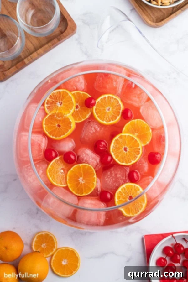 Overhead view of a glass punch bowl filled with sparkling Shirley Temple Party Punch, adorned with floating orange slices and maraschino cherries, ready for serving.